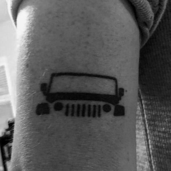 Minimalist black ink tattoo of a Jeep front view silhouette, featuring the iconic grille and headlights, placed on the arm.
