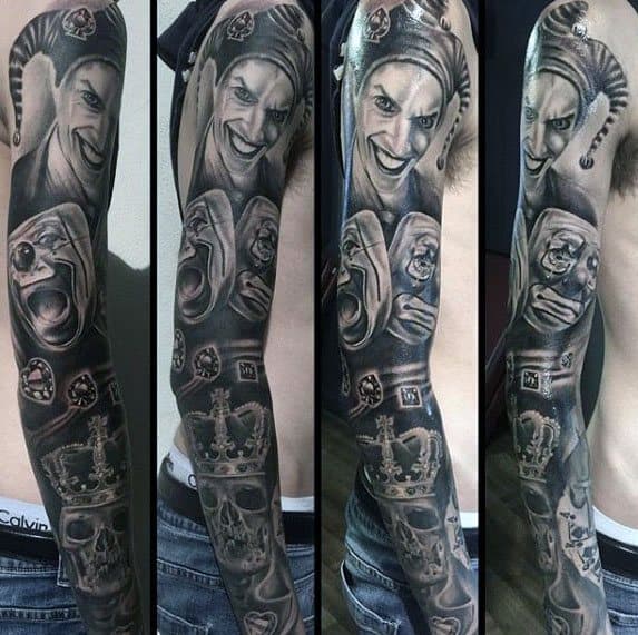 A detailed black and gray sleeve tattoo featuring a jester's face with a mischievous expression, skulls, playing cards, and a crown, creating a dark and playful theme.