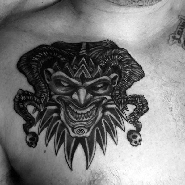 A black and gray tattoo of a sinister jester's face with exaggerated features, adorned with a jagged crown and skull accents, placed on the chest.