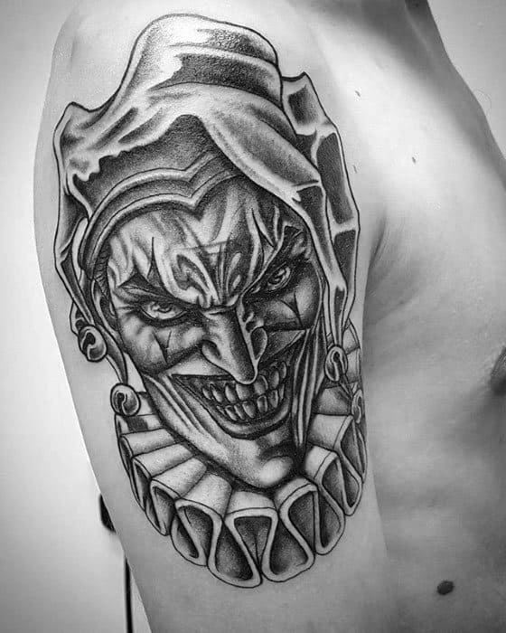 A black and gray tattoo of a sinister jester with a menacing grin, surrounded by a ruffled collar, placed on the upper arm.