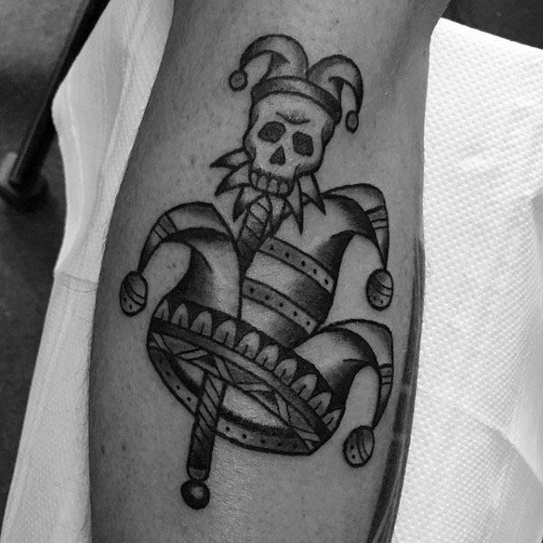 A black and gray tattoo of a jester's hat with a skull at the top, adorned with bells, creating a dark and playful design on the forearm.