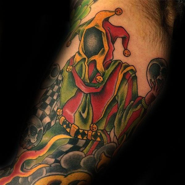 A colorful tattoo of a hooded jester in red and yellow, surrounded by skulls and swirling smoke, placed on the forearm.