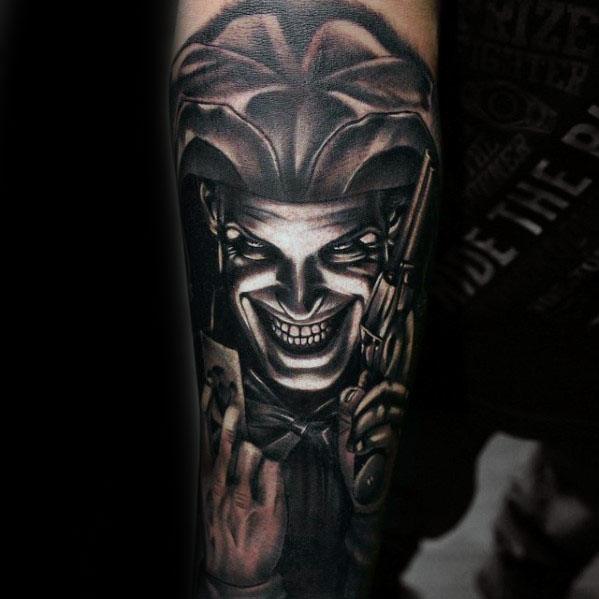A black and gray tattoo of a grinning jester holding a gun and a playing card, with intense features and a menacing expression, placed on the forearm.