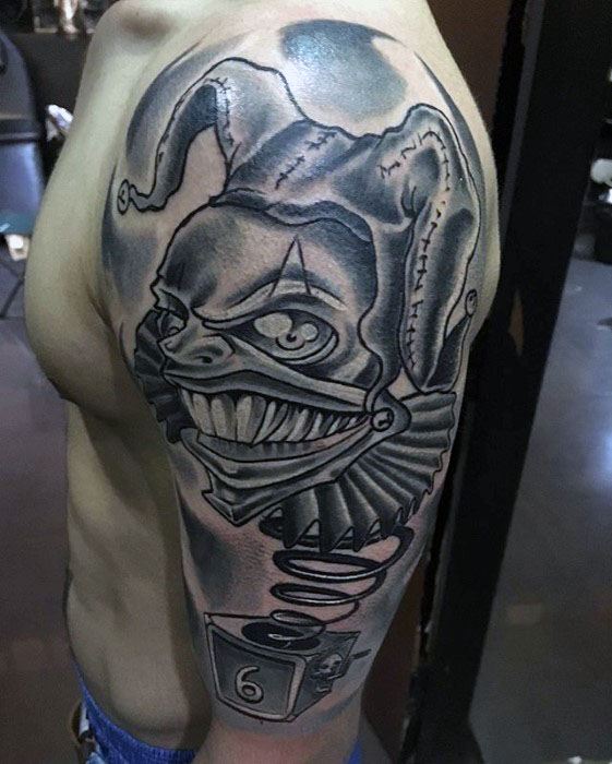 A black and gray tattoo of a sinister jester with a wide grin, accompanied by a spring and a numbered block, placed on the upper arm.