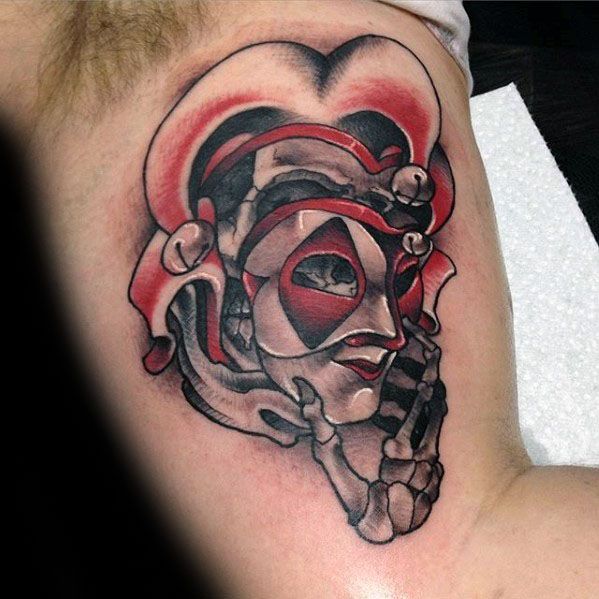 A stylized tattoo of a jester's face, with bold black, white, and red shading, incorporating abstract elements and a skeletal hand design, placed on the upper arm.