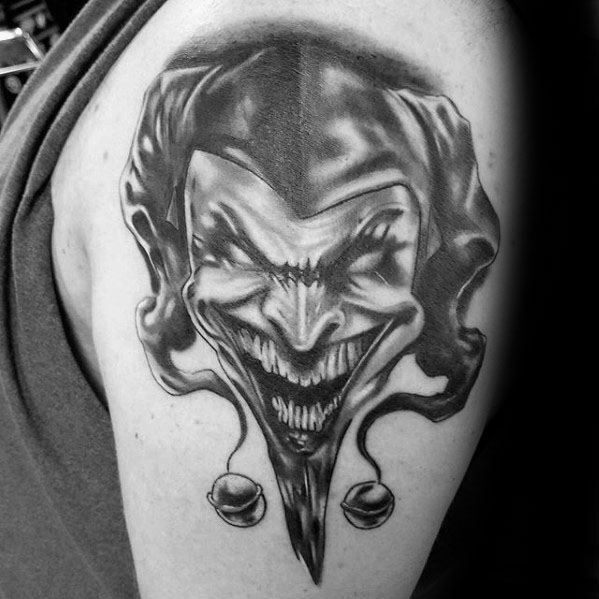 A black and gray tattoo of a menacing jester with an exaggerated grin, showcasing sharp teeth and a dark, intense expression on the upper arm.