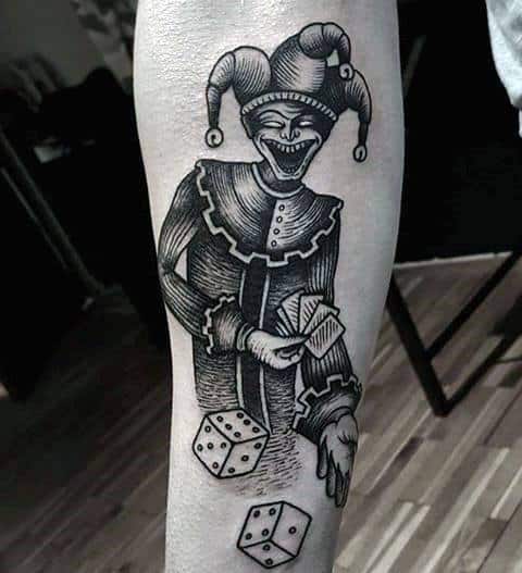 A black and gray tattoo of a jester holding cards with a mischievous grin, accompanied by dice, placed on the forearm.