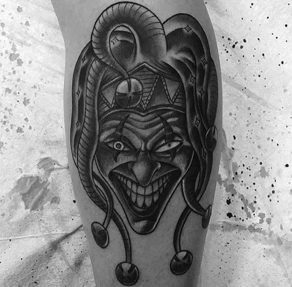 A black and gray tattoo of a sinister jester with exaggerated features and a wide grin, adorned with bells on the jester's hat, placed on the calf.