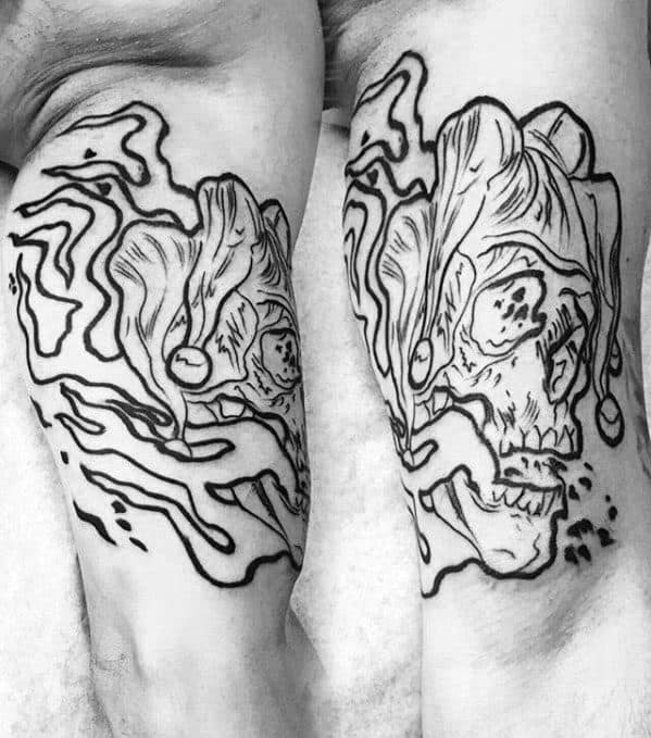 A black outline tattoo of a jester's skull with abstract lines and smoke effects, placed on the forearm, creating a fluid, artistic design.