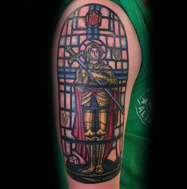 Joan of Arc tattoo in stained glass style on upper arm, featuring golden armor, sword, and vibrant red and pink window-like background.