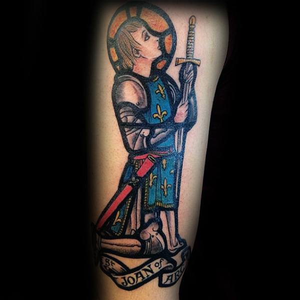 Stained glass-style Joan of Arc tattoo kneeling in prayer, with fleur-de-lis blue robe, sword, and “St. Joan of Arc” banner below.