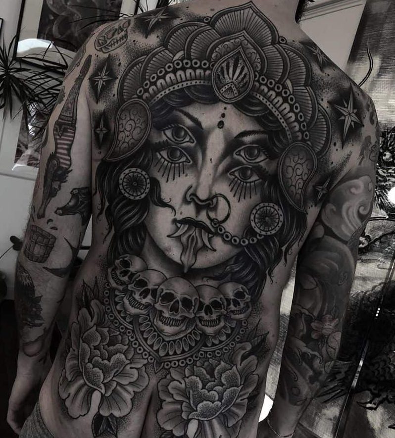 Black and grey Kali tattoo featuring the fierce goddess with four eyes, an ornate headdress, extended tongue, skull necklace, and floral elements