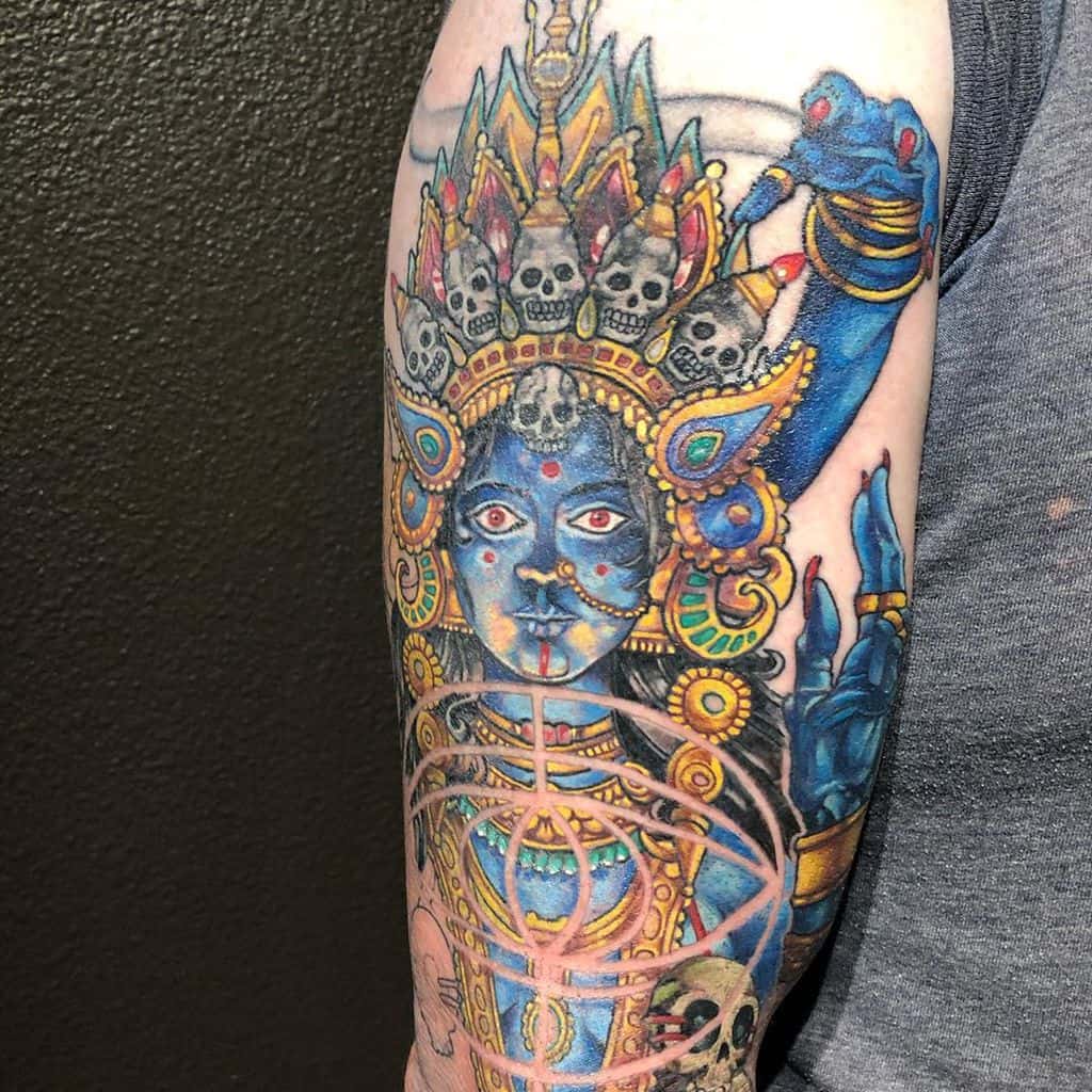 Vibrant tattoo of Kali with skulls above her head, blue skin, intricate patterns, and multiple arms