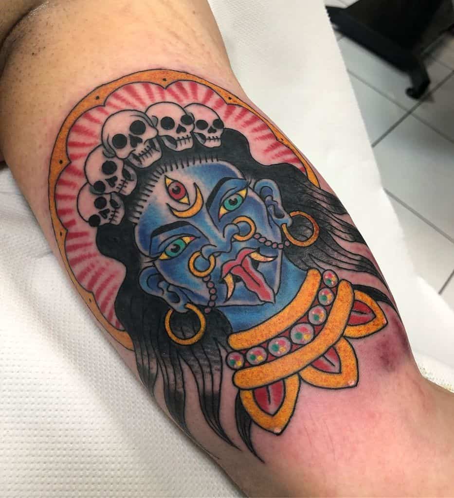 Colorful tattoo of Kali with a third eye, tongue sticking out, adorned with skulls in her hair and a vibrant necklace