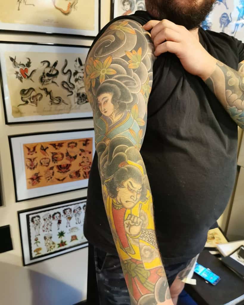 Traditional Japanese-style full-sleeve tattoo featuring a Geisha, a warrior, swirling clouds, and floral patterns in rich colors and bold details.