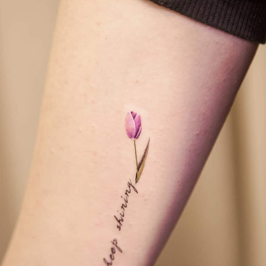 A delicate pink tulip tattoo on the forearm, with a fine green stem and the phrase "keep shining" written in cursive along the design.