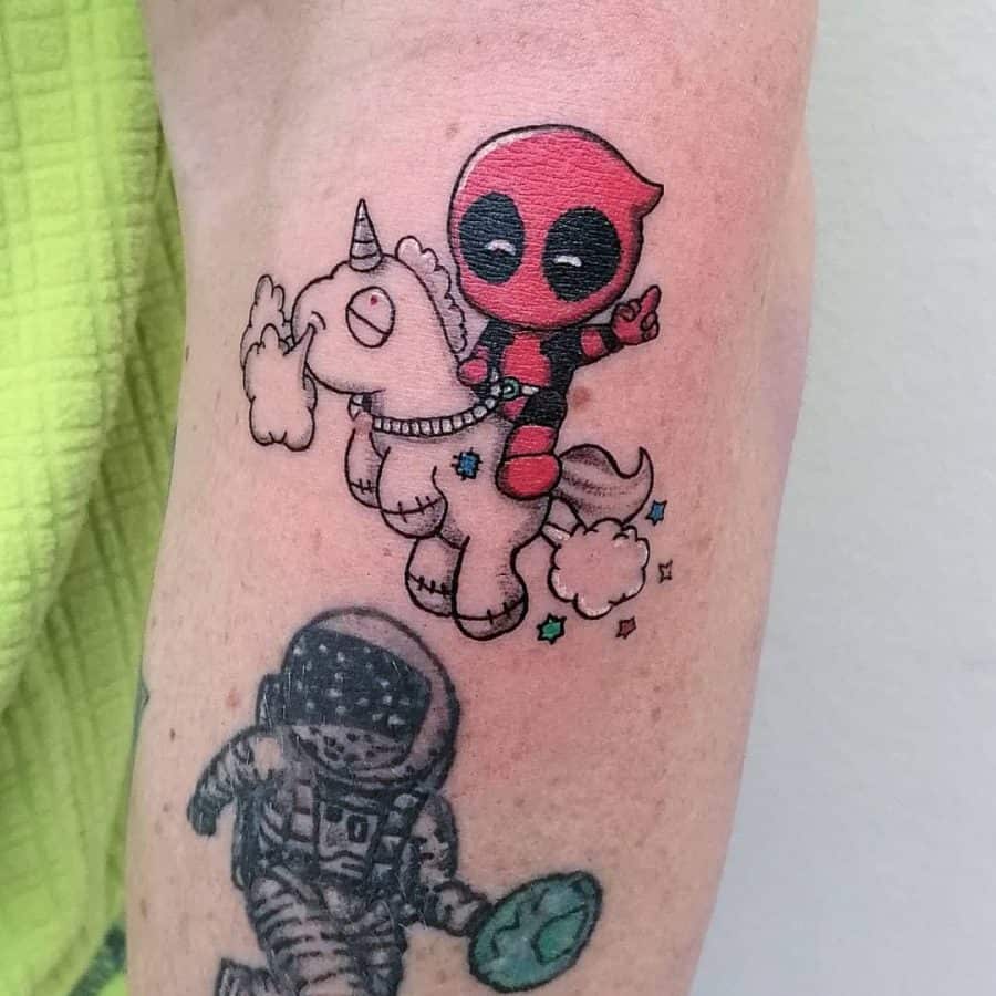 Colorful tattoo of a chibi-style Deadpool riding a cute unicorn, surrounded by clouds and stars, blending humor with fantasy.