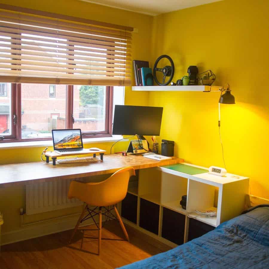 Cozy home office with sunshine yellow walls, a desk, monitors, chair, and a bed in the corner