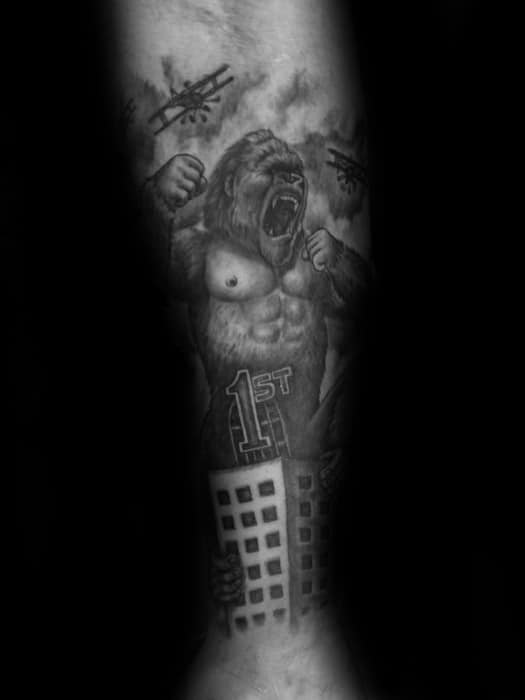 Black and grey King Kong tattoo showing him roaring atop a “1st” labeled skyscraper while biplanes fly around in a stormy sky.