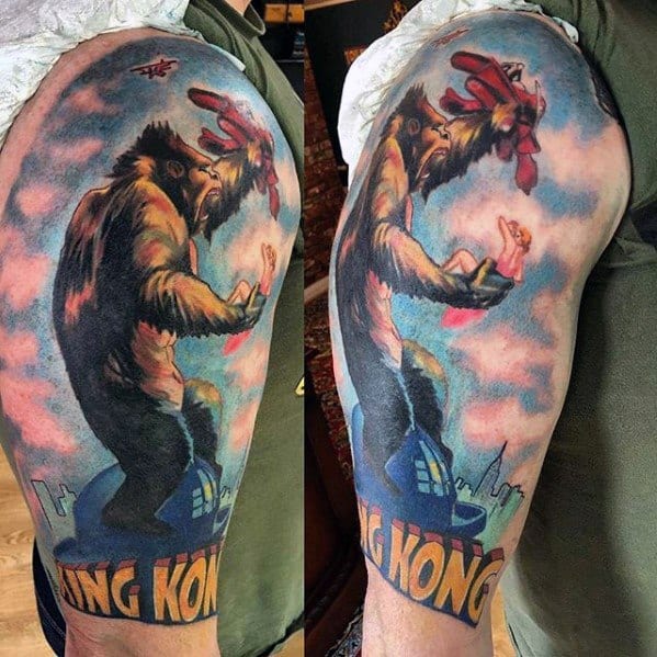 Colorful King Kong tattoo on upper arm featuring Kong atop a building holding a woman, swatting planes, with bold “King Kong” text below.