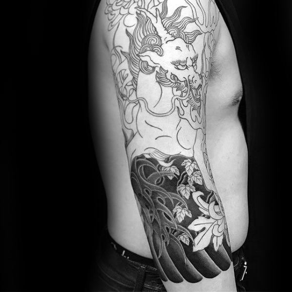 Black and white Kirin tattoo in progress on the upper arm, featuring bold floral patterns and flowing lines blending into dark waves.