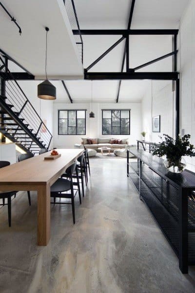 Modern open-plan living room with high ceilings, large windows, a wooden dining table, and metal staircase on concrete floor