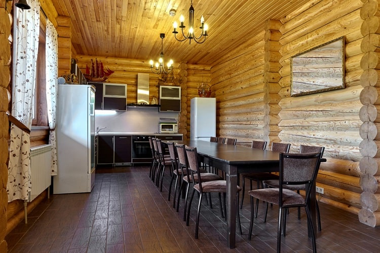 Rustic log cabin kitchen with a long dining table, modern appliances, and wooden walls and ceiling beams