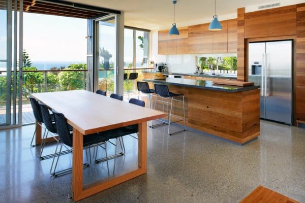 Modern kitchen with wooden accents, concrete floor, a long dining table, hanging lights, and scenic window views