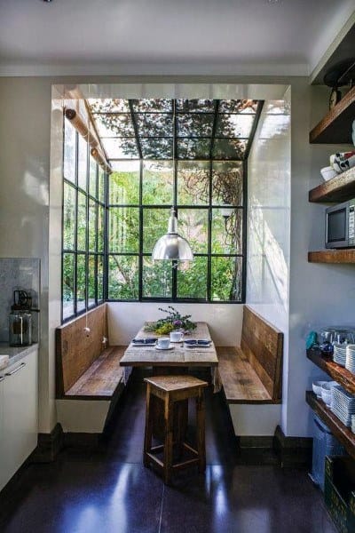 Cozy nook with wooden benches, table, and large windows overlooking a garden, filled with natural light