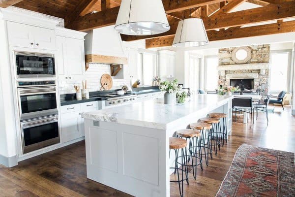 Rustic kitchen with white island, wooden beams, pendant lights, and a stone fireplace.