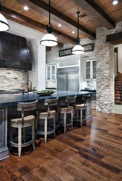 Rustic kitchen with stone walls, dark wood island and floors, pendant lights, and stainless steel fridge
