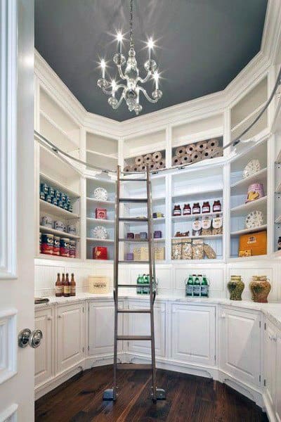 Luxurious kitchen pantry with a built-in ladder, white cabinetry, curved shelving, and a statement chandelier.