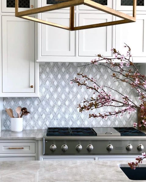 Elegant kitchen with a diamond-patterned stone backsplash, white cabinets, stainless steel stove, and a touch of floral decor