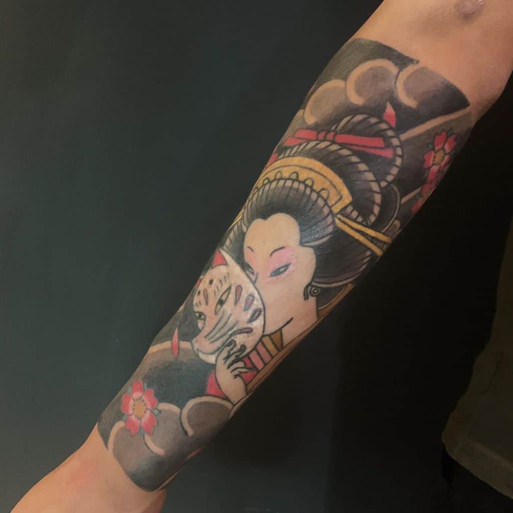 Traditional Japanese-style forearm tattoo of a Geisha holding a kitsune mask, surrounded by swirling clouds, cherry blossoms, and ornate hair accessories.