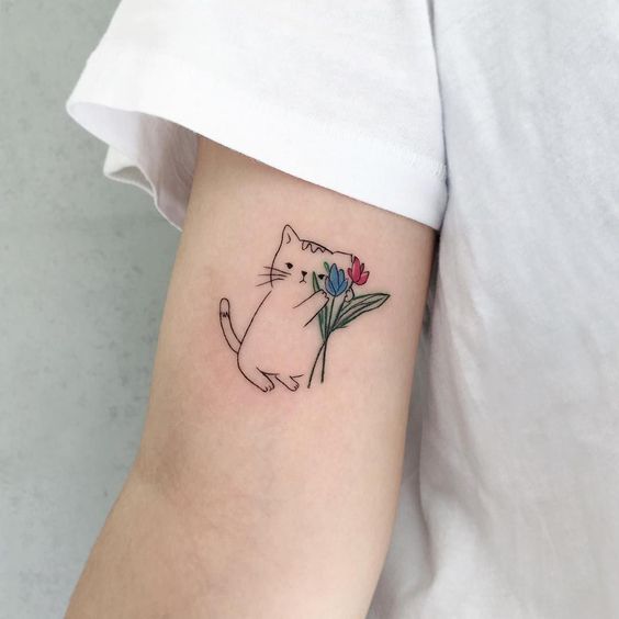 A cute minimalistic tattoo on the upper arm featuring a small cat holding a bouquet of red and blue tulips in a playful, cartoonish style.