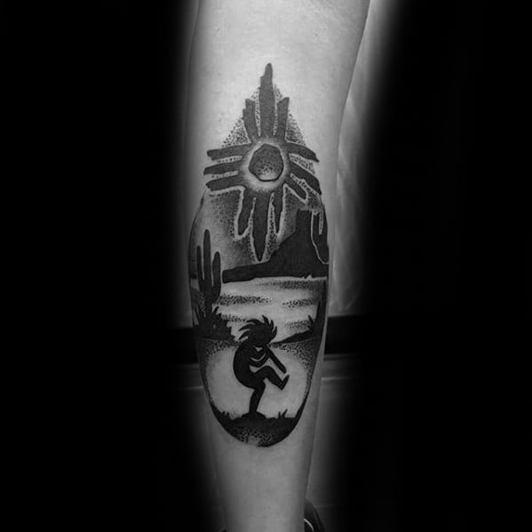 Black and grey Kokopelli tattoo set in a desert landscape with cacti, river, mountains, and a radiant sun symbol above.