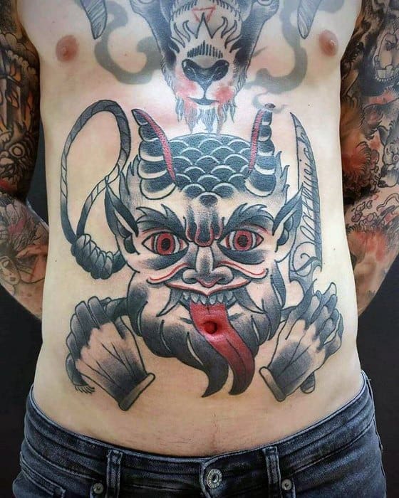 Large Krampus tattoo on stomach showing a grinning demon with red eyes, coiled horns, a red tongue, whips, and clenched fists.