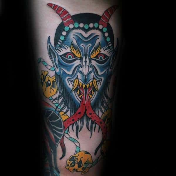 Vibrant Krampus tattoo with a blue demon face, red horns, twin tongues, sharp fangs, and yellow skulls wrapped in green vines.