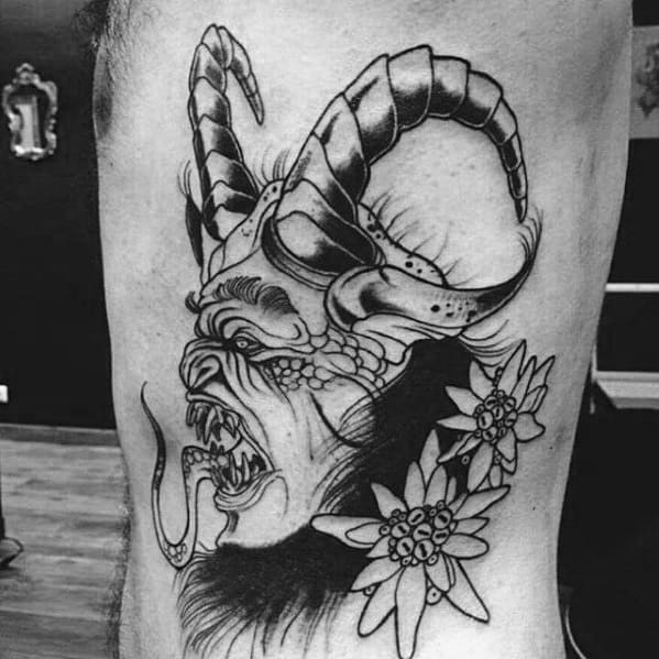 Black linework Krampus tattoo featuring a horned, snarling face with floral accents, long tongue, and dramatic detailing on the side torso.