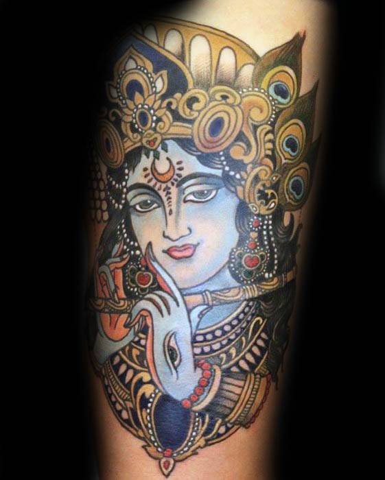 Vibrant Krishna tattoo featuring a blue-skinned Krishna playing the flute, adorned with a richly detailed crown and peacock feathers.