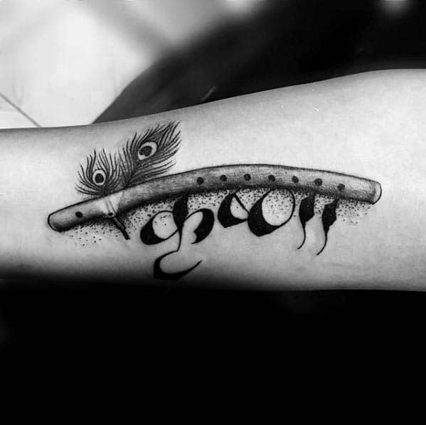 Krishna tattoo with a shaded flute and two peacock feathers above the word "Krishna" written in bold Devanagari script.