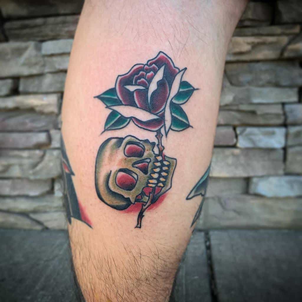 Traditional-style tattoo of a skull with red eyes and a blooming rose, featuring bold lines and vibrant colors on the lower leg.