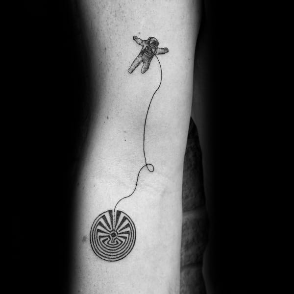 Minimalist astronaut and labyrinth tattoo on a man's arm, featuring an astronaut floating in space, tethered by a thin line to a circular maze.