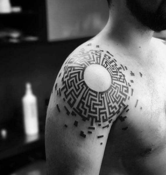 Intricate blackwork labyrinth tattoo on a man's shoulder, featuring a circular maze with a blank center and scattered geometric elements.