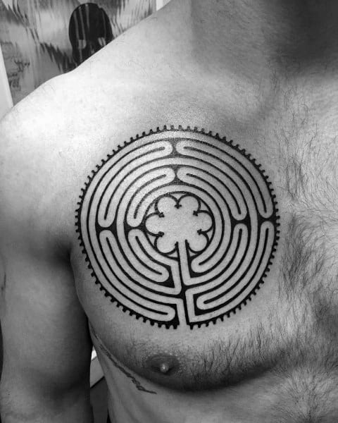 Circular labyrinth tattoo on a man's chest, featuring a bold maze design with a four-leaf clover at the center and a gear-like outer edge.