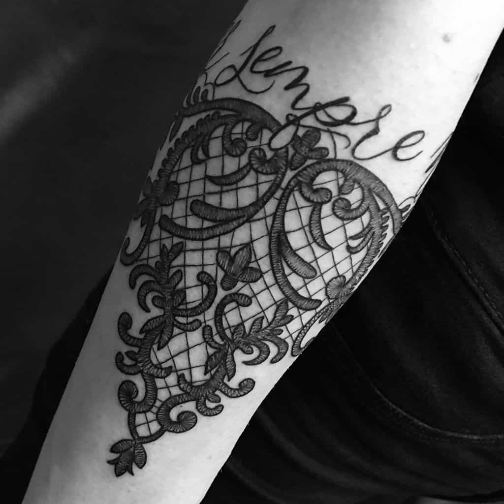 Forearm tattoo featuring an intricately designed black lace heart with ornamental filigree details and delicate script lettering above.