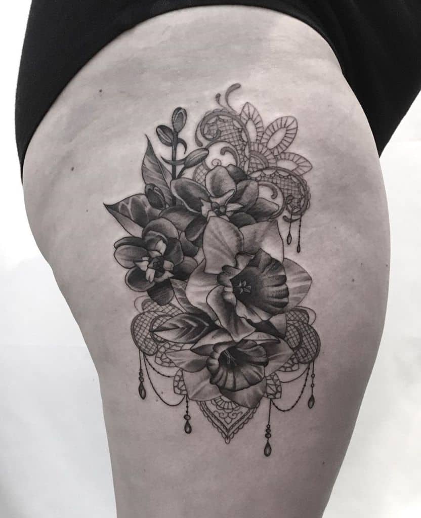 Black and grey thigh tattoo featuring realistic flowers intertwined with intricate lace patterns, ornamental filigree, and delicate beaded chains.