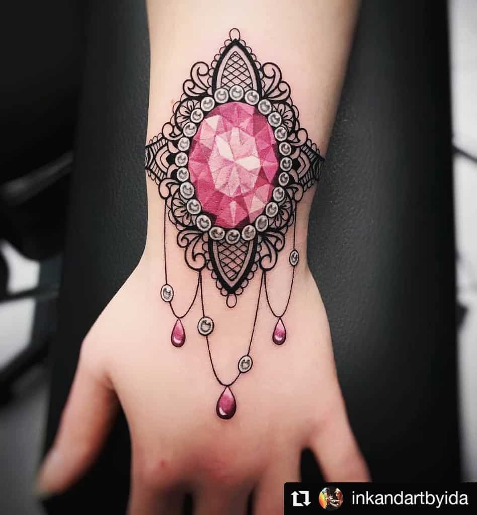 Wrist tattoo featuring an ornate pink gemstone set in intricate black lace with elegant beaded chain details and jewel-like accents.