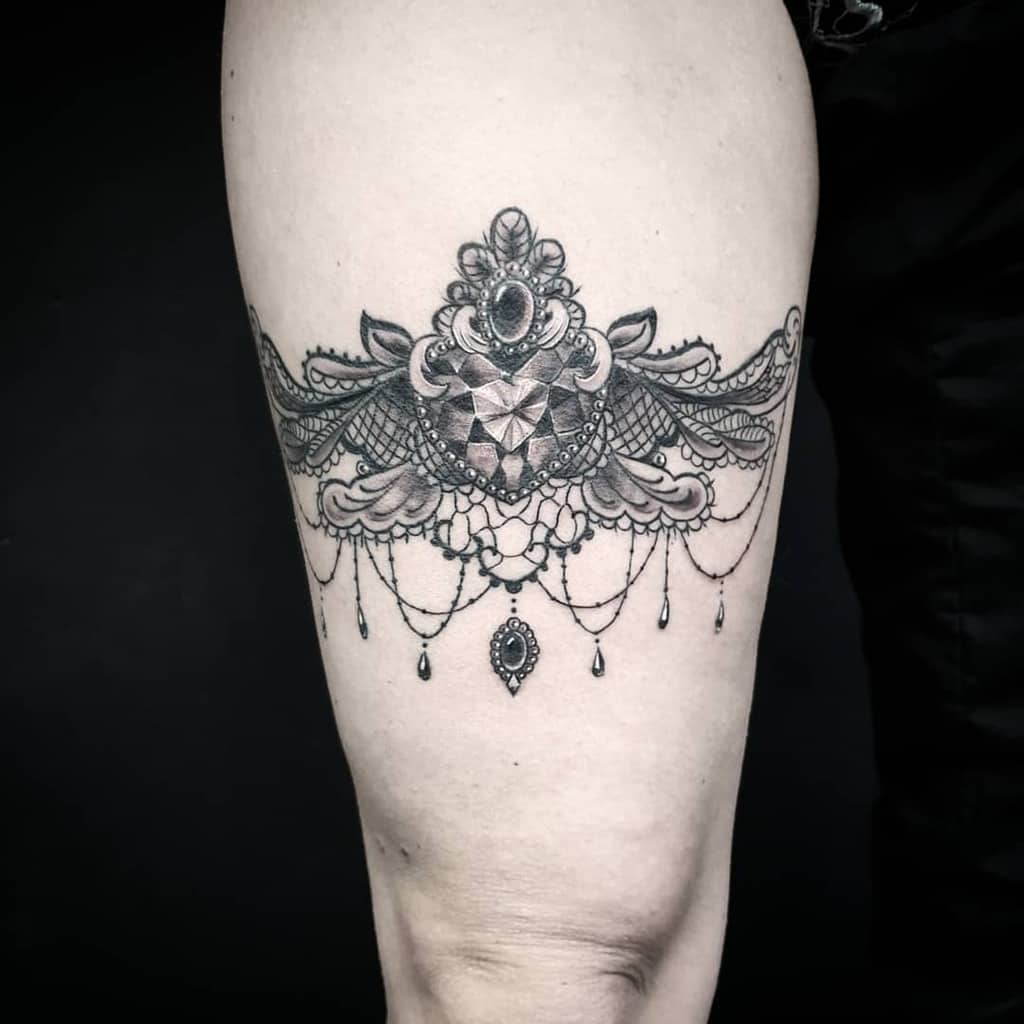 Elegant thigh tattoo resembling a lace garter, featuring a central gemstone, intricate filigree details, and delicate beaded chains.
