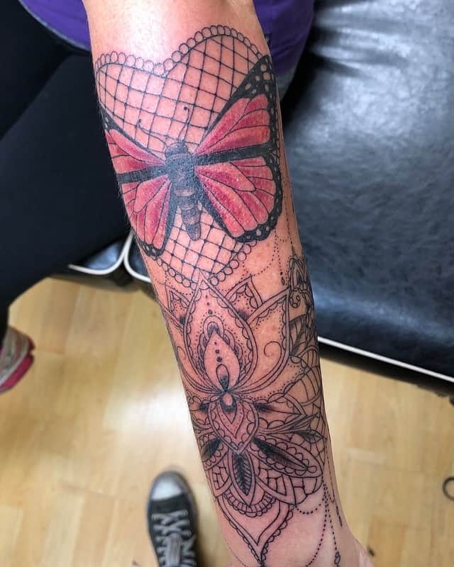 Forearm tattoo featuring a red butterfly with black lace wings, delicate netted patterns, and an ornate mandala-inspired design below.